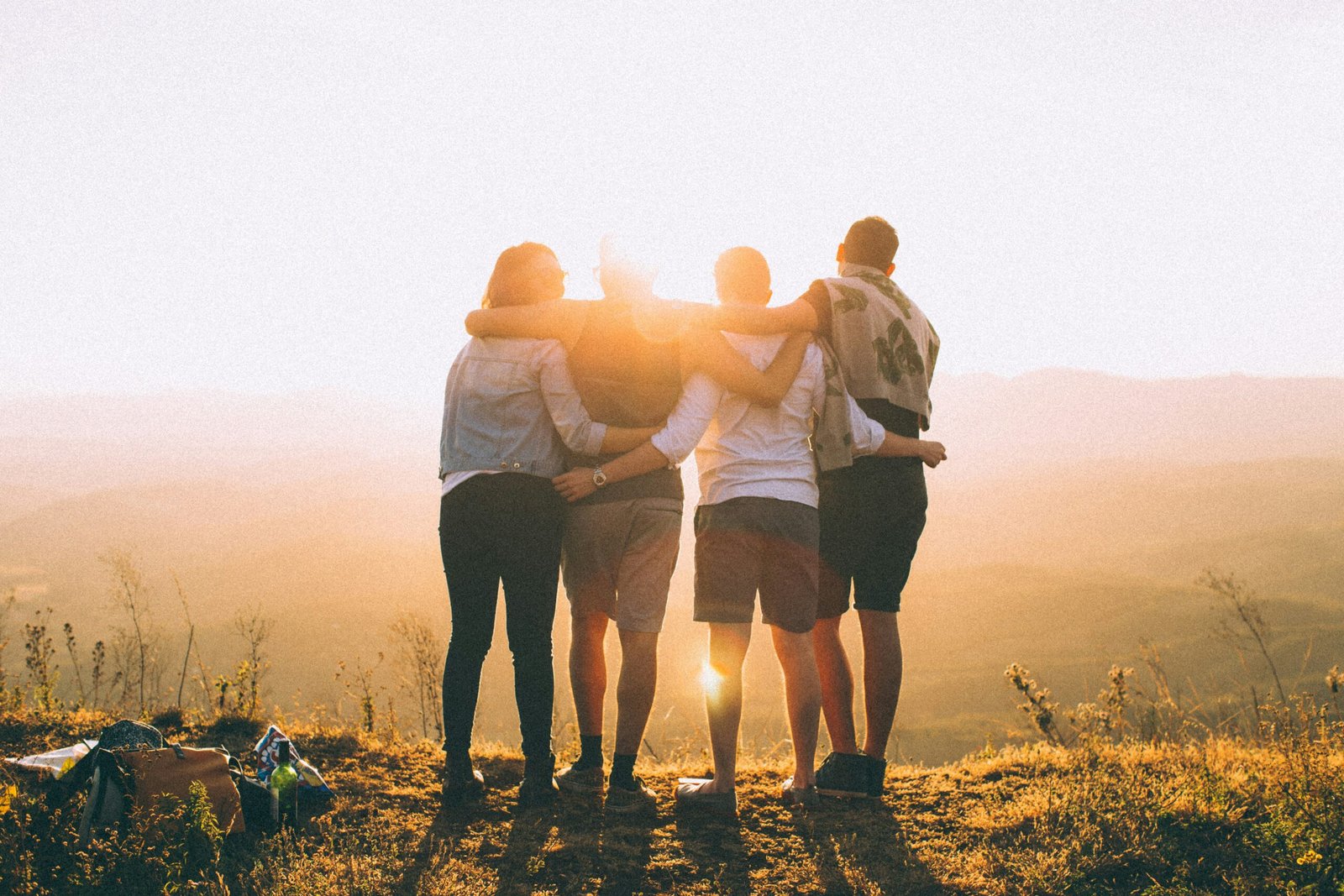 four person hands wrap around shoulders while looking at sunset scaled